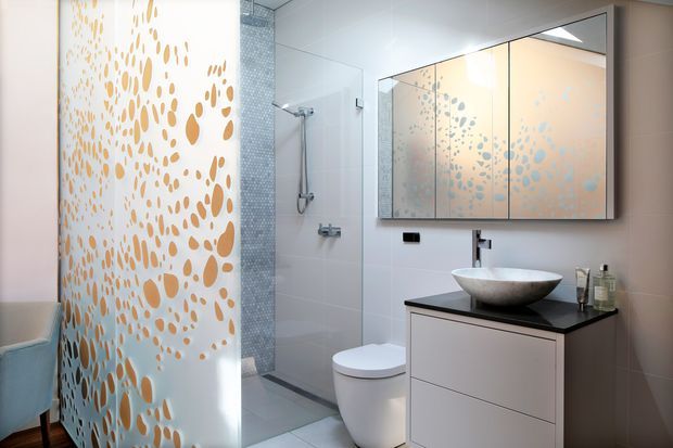 A contemporary bathroom with a frameless glass shower, white wall and floor tiles, a sculptural vessel basin on a dark benchtop, floating pale cabinetry, mirrored wall cabinets, and a decorative translucent screen with organic gold and blue pebble-like patterning. The room is brightly lit with a clean, refined finish.