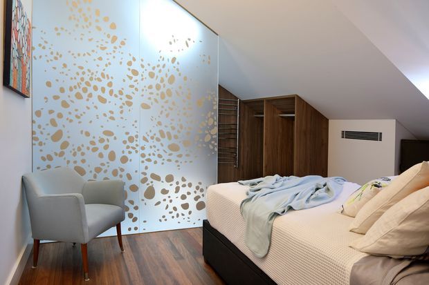 A contemporary bedroom with a low-profile bed, upholstered armchair, warm timber flooring, and extensive built-in timber wardrobe joinery beneath a sloping ceiling. A large decorative translucent glass partition with an irregular gold-toned pattern adds a feature element, while neutral bedding and pale walls create a calm, refined atmosphere.