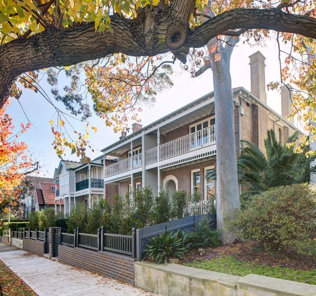 Street-level exterior view of a row of attached brick residences with prominent covered upper-level verandas, white timber balustrades and posts, arched openings, tall chimneys, pitched roofs, and landscaped front gardens behind low brick-and-metal fences. Mature trees frame the scene, conveying an established suburban streetscape.