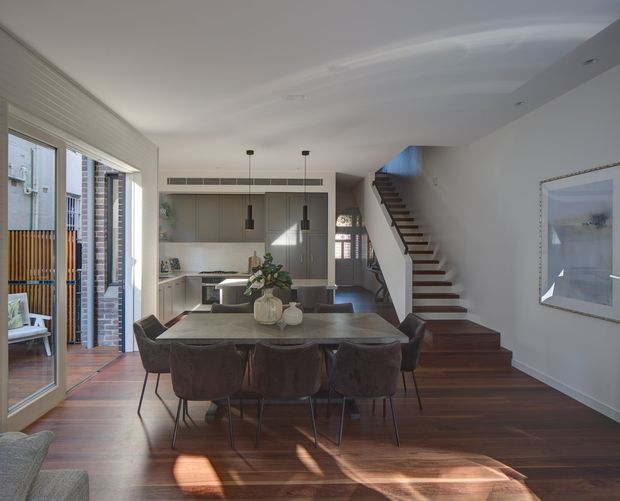 Open-plan residential interior with a dining area in the foreground, a contemporary kitchen at the rear, and a timber staircase rising along the right wall. The room has dark timber flooring, light neutral walls and ceiling, extensive glazing to the left, streamlined grey cabinetry, pendant lighting, and a large dining table with upholstered chairs. A framed artwork and glimpse of living-room seating are also visible.