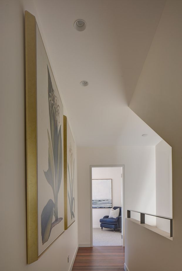 A bright, narrow residential hallway or landing with white walls, recessed ceiling lights, warm timber flooring, and a half-height balustrade overlooking a lower level. Large framed botanical artwork lines the left wall, while an open doorway at the end reveals a furnished sitting area with a blue armchair and coastal artwork.