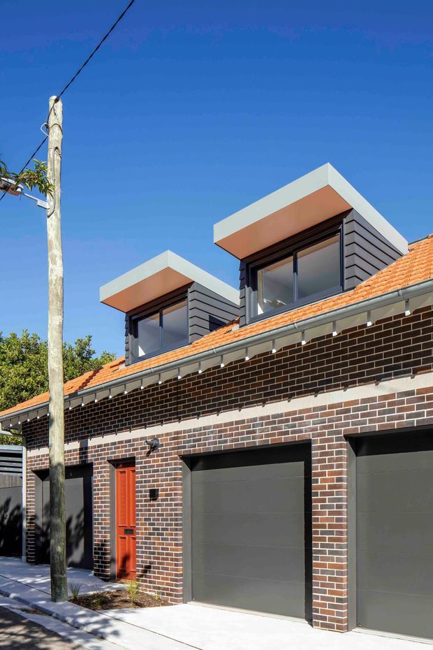 Street-facing exterior of a contemporary multi-level townhouse or residential extension. The facade combines dark brown brickwork, a terracotta tiled pitched roof, charcoal garage doors, and a bright orange-red entry door. Two projecting upper-level dormer-like volumes feature dark horizontal cladding, large windows, and crisp white box roof surrounds. A concrete driveway and narrow landscaped verge are visible in front, with a utility pole at the street edge.