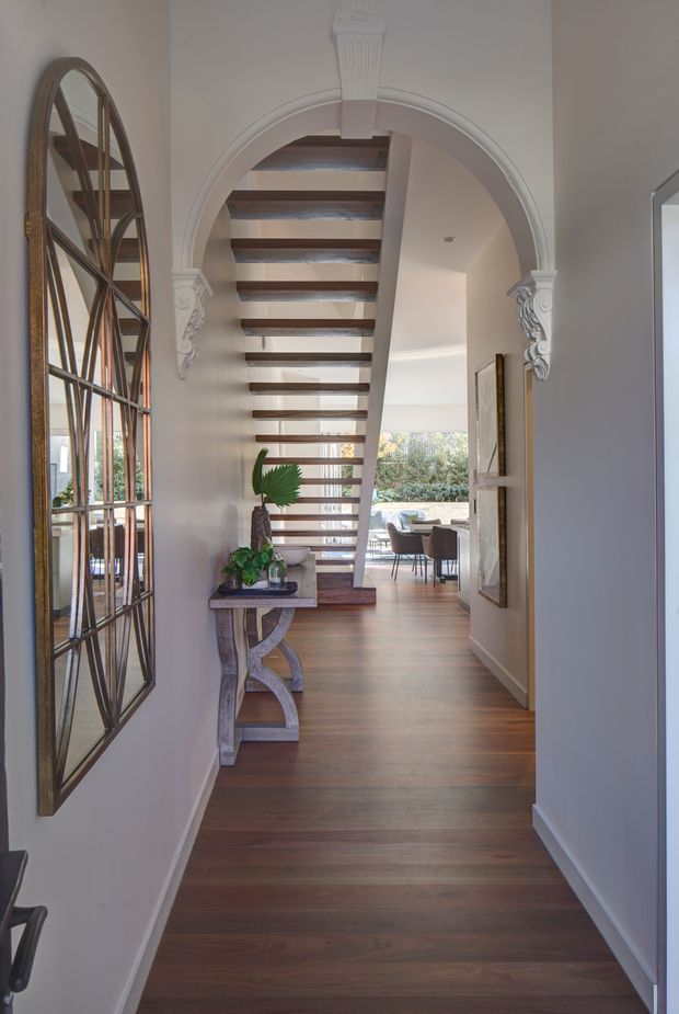 A refined residential hallway viewed toward an ornate arched opening and an open-riser timber staircase. The corridor has warm medium-brown timber flooring, pale painted walls, decorative white arch mouldings with carved brackets, a slim console table with plants, and a large bronze-toned grid mirror. A dining area and garden greenery are visible beyond the staircase.