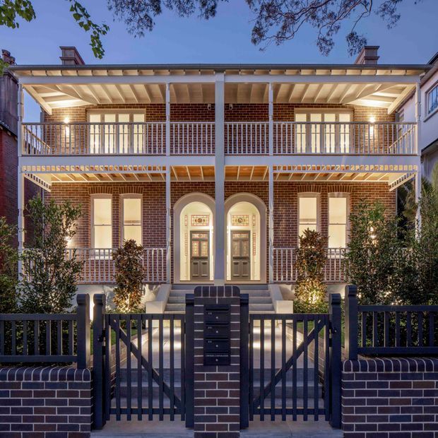 Front exterior view of a symmetrical two-storey brick residence with a prominent covered upper and lower verandah, white timber posts and balustrades, arched entry surrounds, dark framed gates, and landscaped planting. The building is photographed at dusk with warm architectural lighting highlighting the facade.
