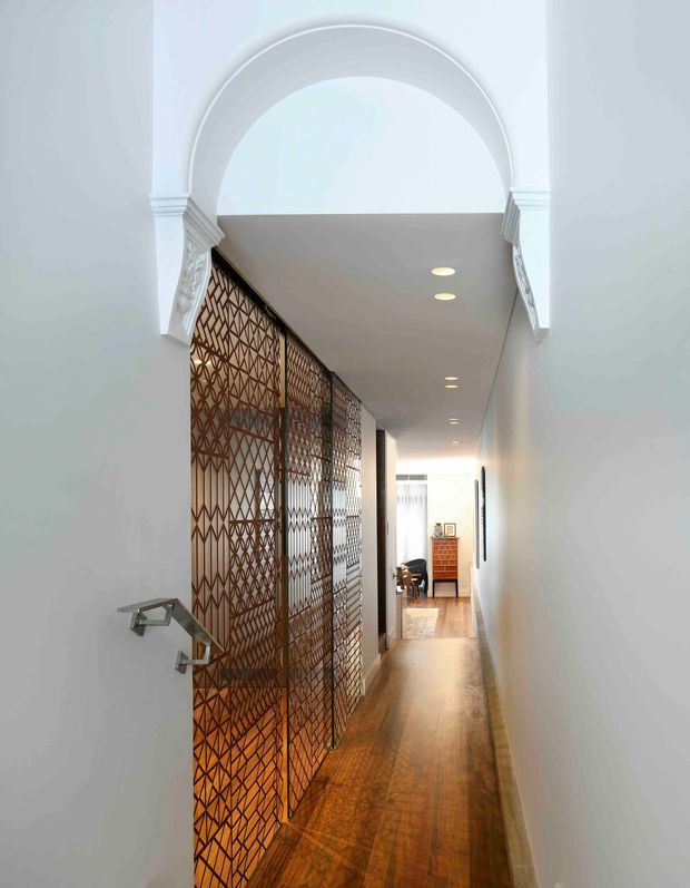 A narrow, bright interior hallway viewed through a decorative arched opening. The corridor has polished warm timber flooring, white walls and ceiling with recessed downlights, and an ornate bronze-toned geometric metal screen enclosing or separating the left side. Decorative classical mouldings frame the arch, while a small dining or living area is visible at the far end.