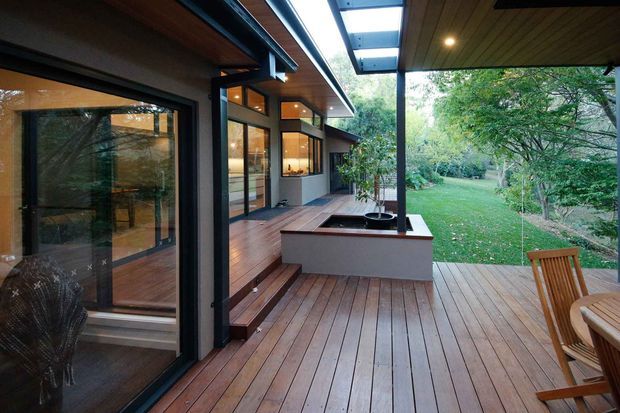 A covered timber deck and outdoor living area beside a modern single-storey home, with dark-framed floor-to-ceiling glazing, warm timber soffits, decking, and a built-in planter. The deck opens onto a leafy garden and includes timber outdoor chairs and a dining setting.