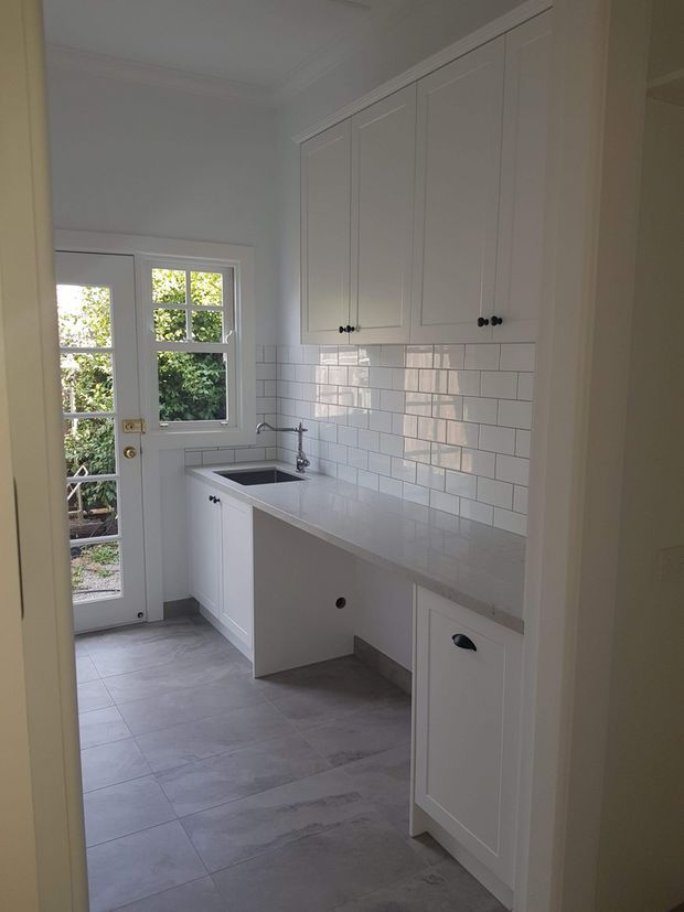 A compact, bright utility/laundry room with white shaker-style cabinetry, a long pale benchtop, inset stainless-steel sink, chrome tapware, and a glossy white subway-tile splashback. Large glazed doors bring in natural light and open to greenery outside. The floor is finished in large grey stone-look tiles, with dark cabinet knobs providing contrast.
