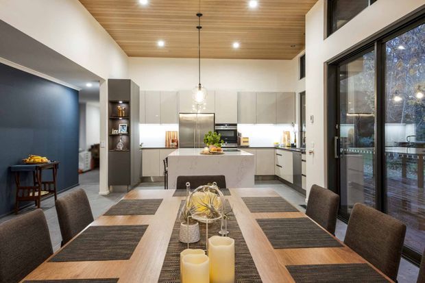A contemporary open-plan kitchen and dining area with a large central island, white cabinetry, stainless-steel appliances, and a timber-lined ceiling. A long timber dining table with upholstered chairs occupies the foreground, while dark blue wall panelling and expansive black-framed glazing add contrast. The space has a tall, airy volume and opens toward an exterior deck or terrace.