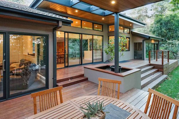 A contemporary timber deck and covered outdoor entertaining area adjoining a modern house in a leafy setting. The space features warm timber decking and soffit lining, dark metal-framed glazing, a raised planter integrated around a structural post, broad steps, and timber outdoor dining furniture. Interior dining and living areas are visible through the glazing.