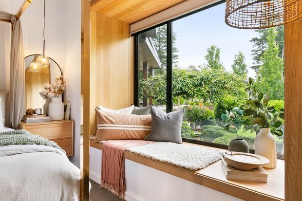 Warm, light-filled bedroom with a built-in timber-framed window seat overlooking a lush garden. The room features pale bedding, soft neutral textiles, a timber bedside cabinet, a large woven pendant light, and extensive timber wall and ceiling finishes.
