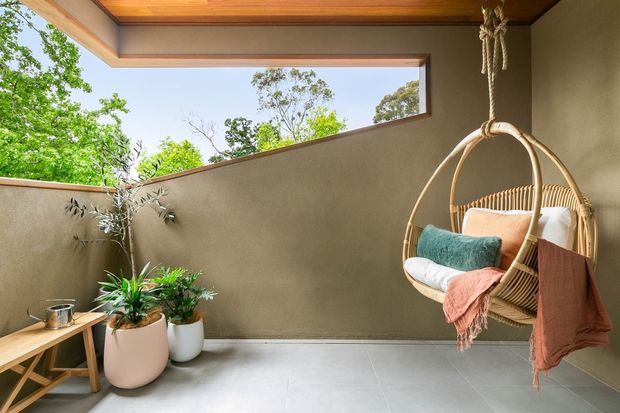 A serene covered outdoor sitting area with warm timber ceiling panels, smooth taupe plaster walls, pale tiled flooring, and a suspended woven hanging chair dressed with cushions and throws. Potted plants and an open clerestory-like wall section bring greenery and daylight into the space.