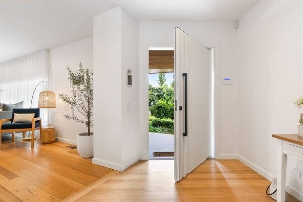Bright contemporary entryway opening directly to an outdoor area, with a large white vertically ribbed pivot door, warm timber flooring, white walls, and a glimpse of an adjacent living area furnished with a lounge chair and floor lamp. Natural greenery is visible beyond the open door.