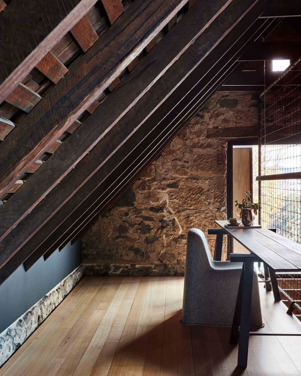 A moody attic-like workspace beneath a steep sloping roof, featuring exposed dark timber rafters, a rough natural stone wall, warm timber flooring, and a long dark desk positioned beside a large glazed opening with metal mesh detailing. A sculptural upholstered chair and a small potted plant complete the compact, rustic workspace.