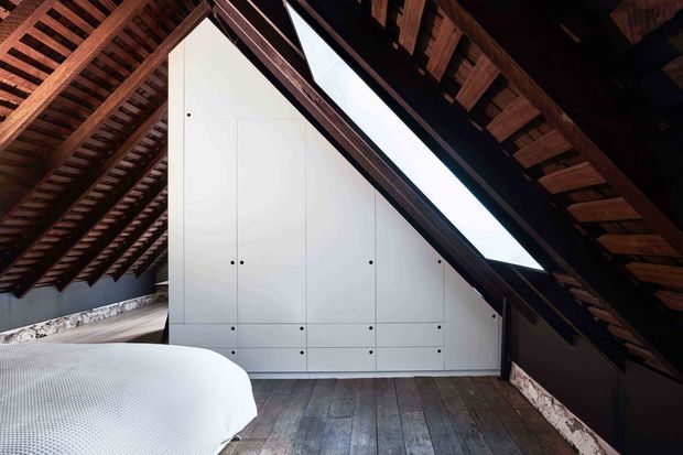 A dramatic attic bedroom beneath a steep pitched roof, featuring exposed dark timber rafters, a large triangular skylight, integrated white built-in cabinetry fitted into the gable wall, and wide weathered timber floorboards. The edge of a bed is visible in the foreground, with a low, warm, rustic architectural character contrasted by crisp contemporary joinery.