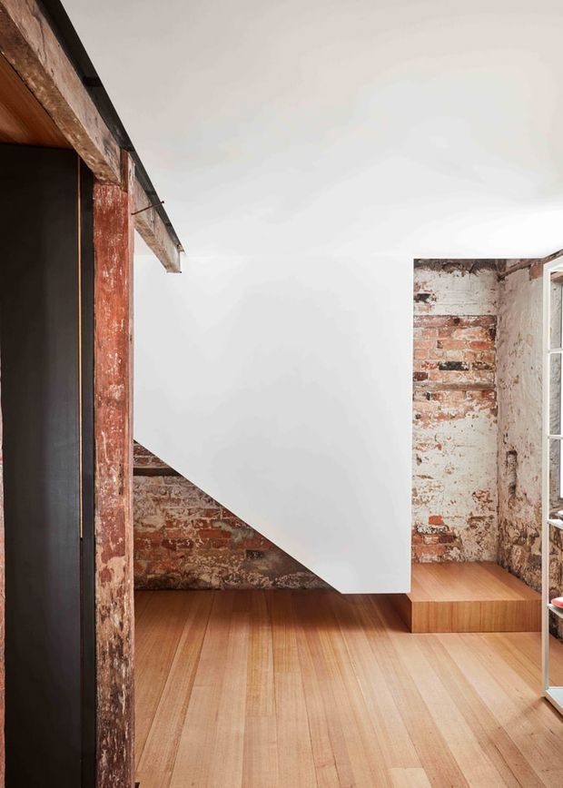 Interior architectural photograph of a compact loft-like room with exposed, weathered red brick walls and timber framing, warm timber plank flooring, and a sharply sculptural white stair or mezzanine enclosure. A black built-in element is visible at left, with a window and open shelving at right. The space has a raw adaptive-reuse character balanced by clean contemporary detailing.
