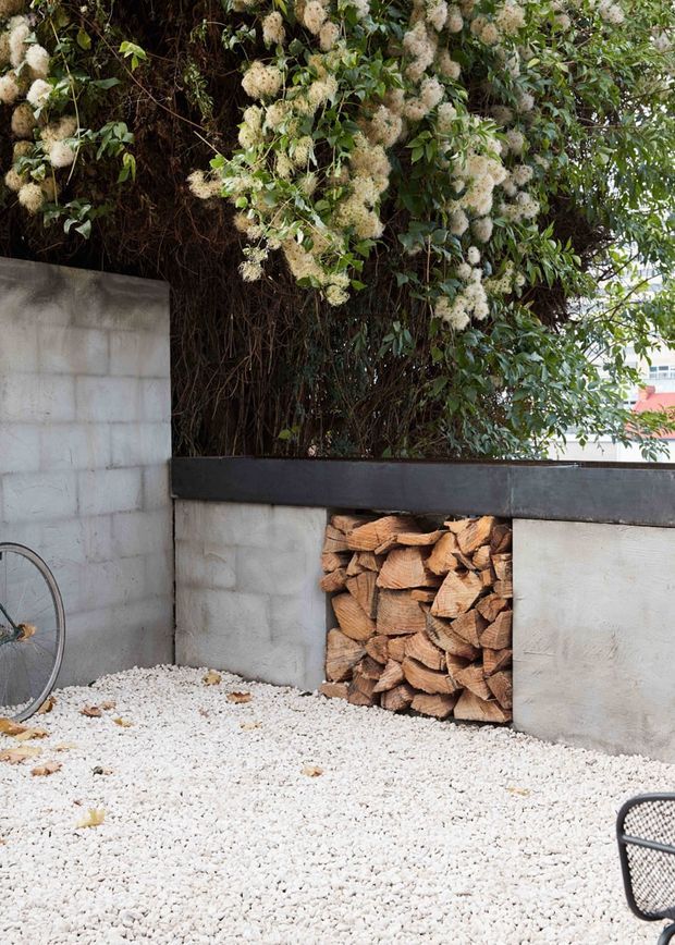 A small landscaped courtyard or outdoor area with white gravel flooring, raw concrete block walls, a dark horizontal ledge, and a built-in firewood storage niche filled with split logs. Dense flowering greenery rises above the wall, with partial bicycle and outdoor furniture visible at the edges.