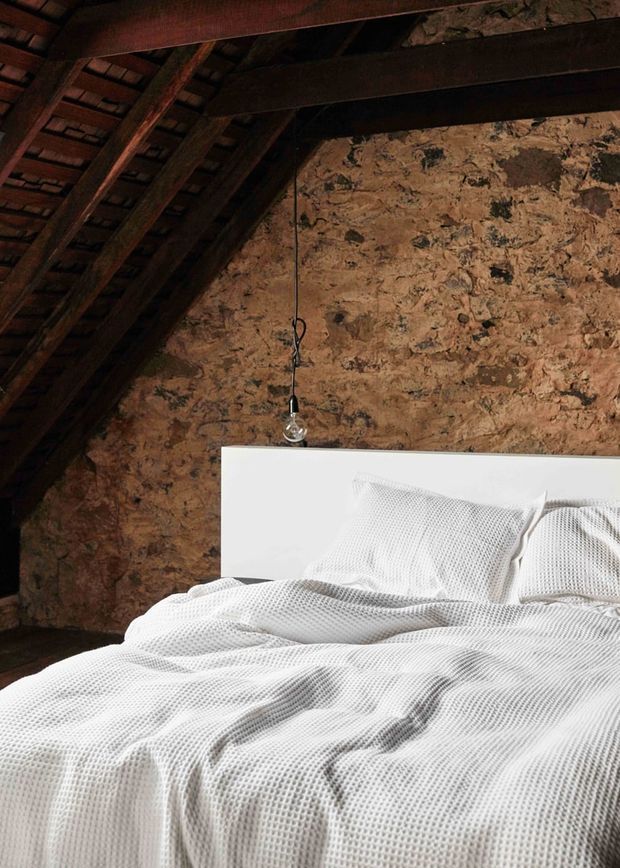 A rustic bedroom beneath a steeply pitched roof, featuring exposed dark timber rafters and beams, rough natural earthen-textured wall surfaces, and a simple white bed with textured white bedding. A single hanging pendant light adds a minimal contemporary contrast to the rugged structure.