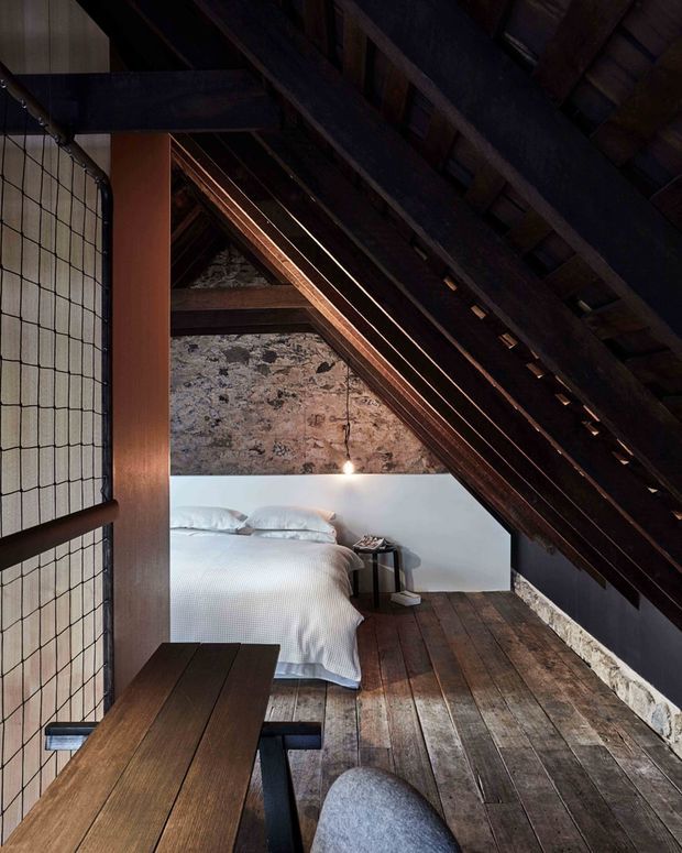 A dark, atmospheric loft bedroom tucked beneath a steeply pitched timber roof. Exposed dark rafters and structural beams frame a weathered stone wall, while a white bed sits against a simple white half-height wall. Wide reclaimed timber floorboards, a rustic timber table, a small bedside table, and a large grid-patterned window create a warm, rustic retreat.