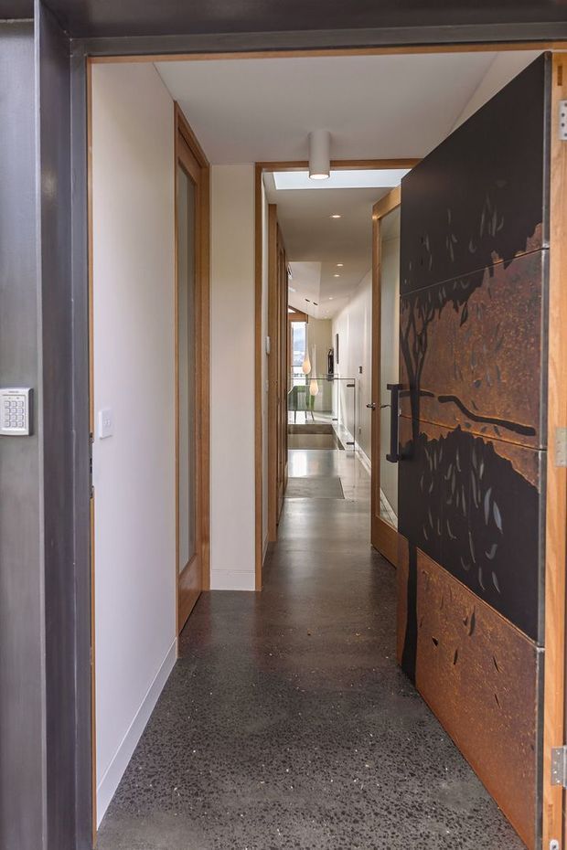 A narrow, modern interior hallway viewed through an open entrance door. The corridor has polished dark exposed-aggregate concrete flooring, white plaster walls, warm timber door frames and cabinetry, recessed ceiling lights, and a large sculptural rusted metal door or screen with abstract cut-out detailing. A glass balustrade and bright stair or landing area are visible at the far end.