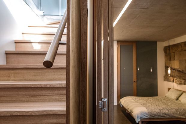 Contemporary interior featuring a timber staircase with a cylindrical wooden handrail and vertical timber slat partition. The view also includes a partially visible bedroom with a low bed, neutral bedding, timber-framed door, dark wall, and exposed natural stone feature wall. A linear recessed ceiling light and skylight add modern architectural detailing.
