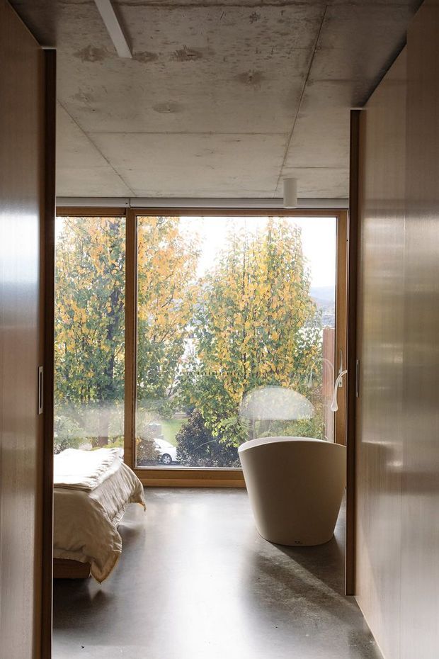 A compact, minimalist bedroom with a freestanding bathtub positioned beside expansive timber-framed glazing. The room features floor-to-ceiling built-in timber cabinetry, a raw exposed concrete ceiling, polished concrete flooring, and a restrained neutral palette. Autumn foliage is visible outside through the large window.