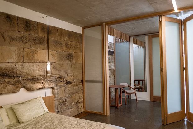 A warm, compact bedroom with a bed in the foreground and an exposed rough stone feature wall. Timber-framed translucent sliding or folding screens open toward a small adjoining sitting area with wooden chairs and a table. The space has a low raw concrete ceiling, dark polished-looking flooring, and simple pendant lighting, creating a rustic yet refined architectural interior.