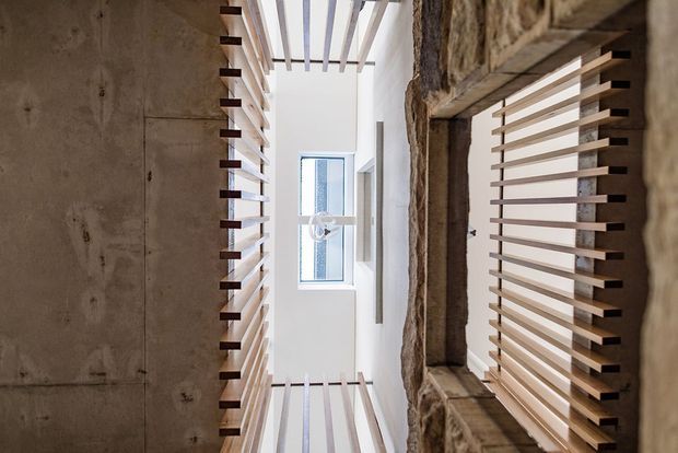 A narrow, vertically oriented architectural interior view showing a high open void or stairwell. Rough exposed masonry and concrete walls frame white horizontal timber slats and a skylit opening, creating a strong contrast between raw structural surfaces and light screening elements.
