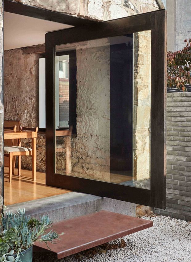 Architectural detail of a dark-framed glazed pivot door set within a rough stone and masonry wall. The open doorway connects a polished timber-floored dining area to a small gravel courtyard, with a timber dining table and chairs visible inside. Strong material contrast between dark metal framing, textured stone, concrete steps, reddish paving, and pale gravel.