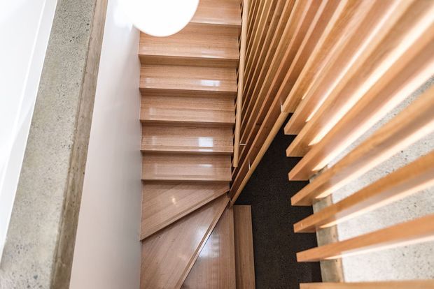 A modern interior stairway viewed from above, featuring warm timber treads and matching timber slatted screening along the side. The stairs are bordered by smooth white walls, a raw concrete structural element, and a dark contrasting stairwell surface. A round white ceiling light is partially visible.