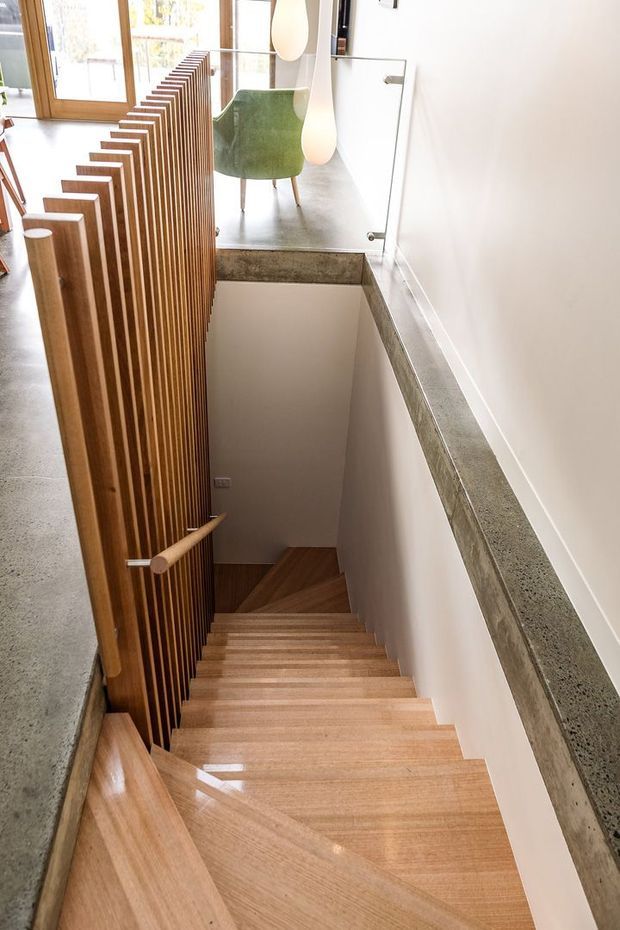 Bright contemporary interior view looking down a timber staircase. The stair has warm wood treads and handrail, a vertical timber slat balustrade, white walls, and a grey polished concrete or stone landing edge. A green upholstered chair and large windows are visible at the upper level.