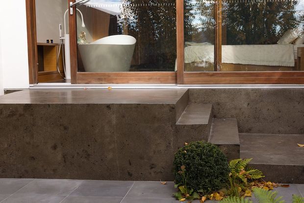 Exterior view of a contemporary home with a sunken concrete courtyard or terrace. Large timber-framed glazing reveals a freestanding bathtub and bedroom interior, while broad concrete steps and a raised concrete retaining wall define the space. Grey tiled paving and integrated landscaping complete the minimalist setting.