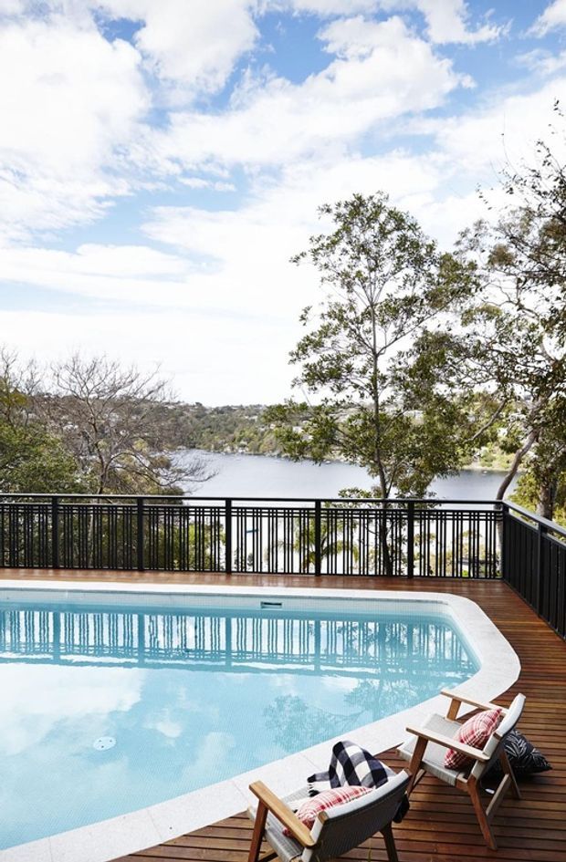 A bright outdoor pool terrace overlooking a calm wooded body of water. The rectangular swimming pool has pale stone coping, a dark timber deck, black metal balustrades, and timber-framed lounge chairs with patterned cushions. Mature trees and a partly cloudy blue sky form the background.