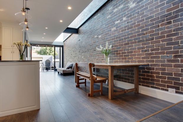 Open-plan contemporary interior combining a kitchen, dining area and living space. A long exposed brick feature wall runs alongside dark timber-look flooring, with a simple timber dining table and bench seating. White kitchen cabinetry, roof glazing or a high clerestory opening, black-framed glazing and pendant lights create a bright, warm space.