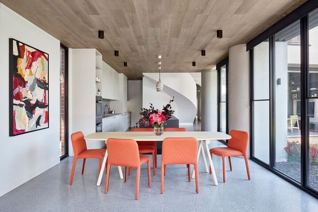 A bright contemporary open-plan dining area beside a compact kitchen, with a long white dining table surrounded by coral upholstered chairs. The room features full-height black-framed glazing, a light timber-clad ceiling, pale cabinetry, polished grey flooring, and a curved white feature wall. An abstract artwork and floral arrangement add colour.