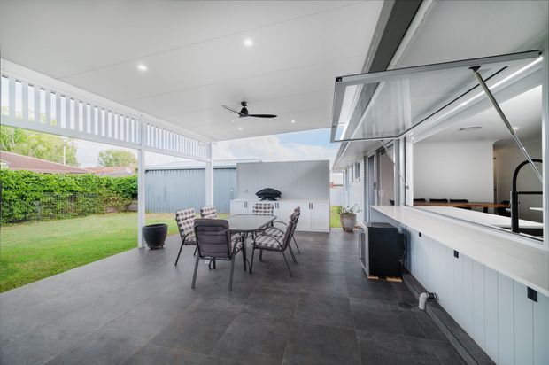 Covered outdoor entertaining area with a dining table and chairs, ceiling fan, tiled floor, outdoor kitchen cabinetry and barbecue, and a large servery window opening to the interior. The space overlooks a lawn and garden and has a bright, contemporary feel.