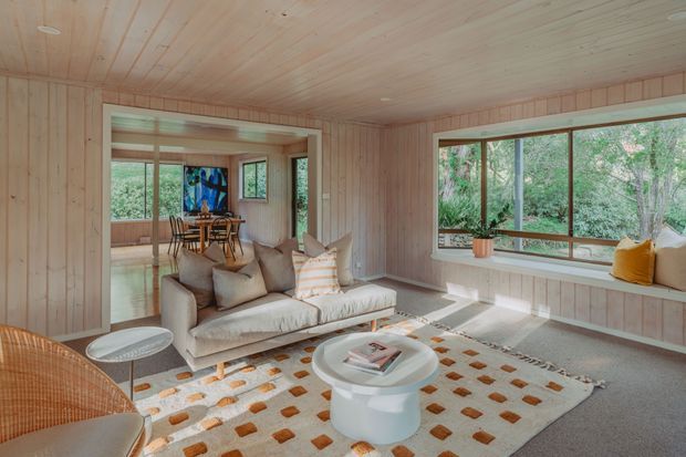 Warm, light-filled living room clad in pale timber panelling, with a large upholstered sofa, sculptural white coffee table, patterned rug, woven occasional chair, and extensive windows overlooking greenery. An open connection leads to a dining area visible through the room.