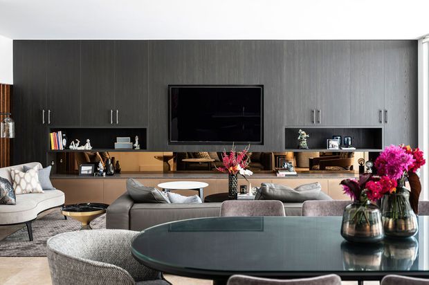 Contemporary open-plan living and dining area featuring a dark timber entertainment wall with integrated cabinetry, shelving and wall-mounted television. A large curved glass dining table, upholstered dining chairs, sectional seating, sculptural coffee table and floral arrangements create a refined, layered interior.