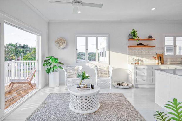 Bright open-plan coastal living area with a compact white kitchen, pale timber-look flooring, woven lounge furniture, a round coffee table, textured rug, indoor plants, and direct access to a small timber deck through wide glazed doors. The room is filled with natural light and has a ceiling fan, white cabinetry, open timber shelving, and a white brick-pattern splashback.