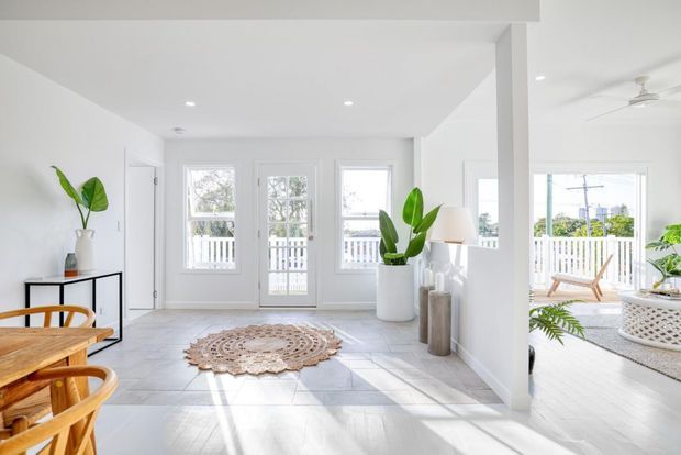 Bright, airy coastal-style open-plan living space with white walls, pale tiled flooring, timber furniture, tropical plants, and abundant glazing opening onto a white-railed balcony or terrace. A woven circular rug and natural materials add warmth, while a ceiling fan and outdoor seating reinforce the relaxed resort-like atmosphere.