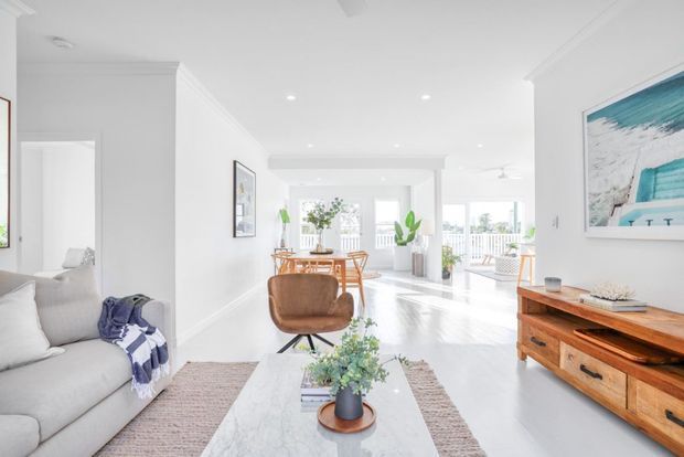 Bright open-plan coastal-style living and dining area with white walls, light flooring, large windows, a pale sectional sofa, timber dining furniture, a marble-look coffee table, and abundant indoor plants. A balcony or exterior view is visible through the glazing.