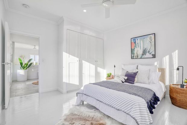 Bright, airy white bedroom with a large upholstered bed dressed in layered white and blue-grey striped bedding, built-in panelled wardrobes, framed botanical artwork, a woven basket, and a ceiling fan. An open doorway reveals an adjoining bathroom, while strong sunlight creates patterned shadows across the room.