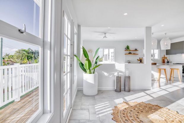 Bright open-plan residential interior with white walls, large white-framed windows and glazed doors, pale tiled flooring, and strong natural sunlight. A living area with a white sofa and decorative shelving opens directly to a contemporary kitchen with grey cabinetry, white subway-tile splashback, and a timber breakfast bar. A balcony with white balustrades is visible through the glazing.