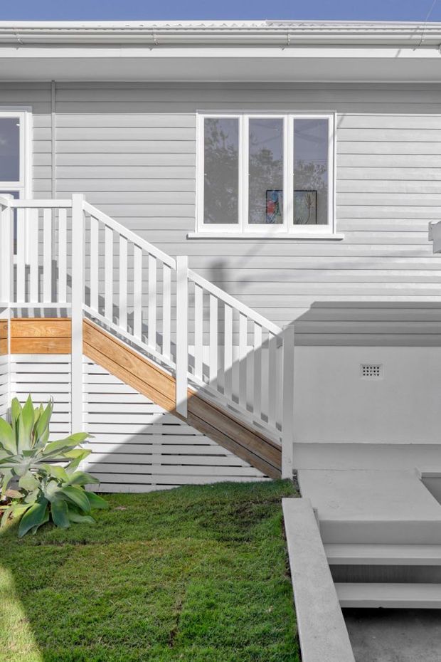 Exterior view of a renovated light-grey weatherboard house, featuring white-framed windows, gutters and railings, with a timber-accented staircase leading to an elevated entry deck. A concrete stairwell descends beside a small grassed garden area.
