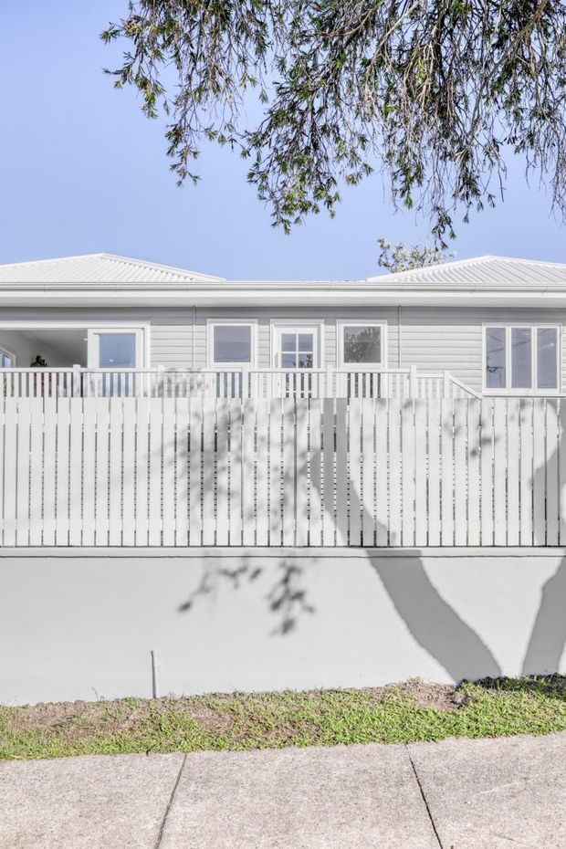 Bright exterior view of a single-storey weatherboard building with a pale metal gabled roof, white-framed windows, and a raised white timber balcony or verandah enclosed by a vertical picket privacy screen. The facade is photographed from across a concrete footpath, with strong tree shadows cast across the wall and screen.