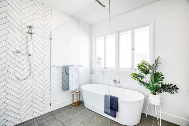 Bright contemporary bathroom featuring a freestanding white bathtub beside a frameless glass shower enclosure. The shower wall is clad in white herringbone tiles, while the floor uses large dark grey tiles. White-framed windows provide abundant natural light, and a potted palm adds a small natural accent.