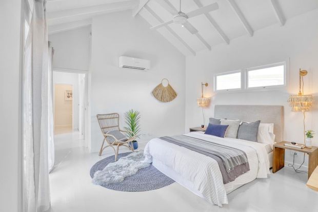 Bright, airy bedroom with a white coastal-inspired interior, a large bed dressed in layered white and grey bedding, woven rattan armchair, textured circular rugs, natural fibre wall decor, pendant lights, and exposed white ceiling beams with a ceiling fan. High-level windows and white finishes create a serene, spacious feel.