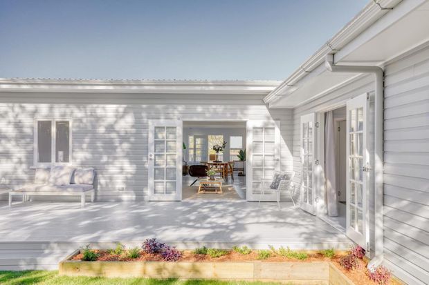 Bright, white-painted weatherboard courtyard or patio area connecting to an open-plan interior through glazed French doors. The space features a pale timber deck, raised garden beds, white-framed windows and doors, and a covered porch with traditional eaves and gutters. Dining furniture is visible inside, with strong natural light and leafy shadows creating an airy residential setting.
