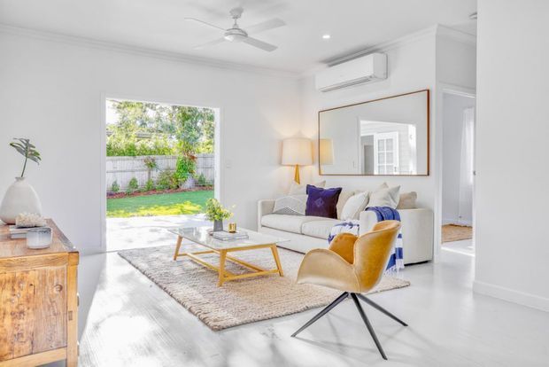 Bright, airy living room with white walls and white-painted timber flooring, a large opening to a leafy outdoor area, a cream sofa, mustard lounge chair, patterned rug, timber coffee table, sideboard, wall mirror, ceiling fan, and wall-mounted air conditioner. The room has a clean, contemporary coastal feel with restrained colourful accents.