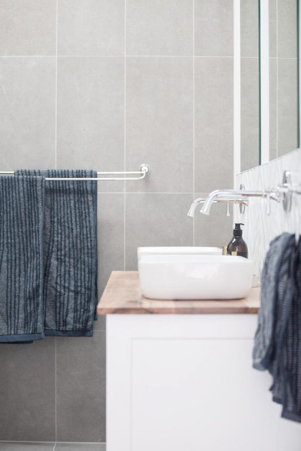 A bright, compact bathroom featuring large light-grey wall tiles, a white vessel basin on a timber benchtop, white vanity cabinetry, chrome wall-mounted taps, a large mirror, and dark charcoal towels on simple rails.