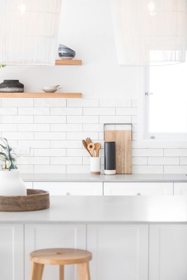 Bright, minimalist white kitchen with a subway-tile backsplash, white shaker-style cabinetry, pale countertop, open timber shelves, and a white-framed window. Natural timber accessories and a simple stool add warmth to the predominantly white space.